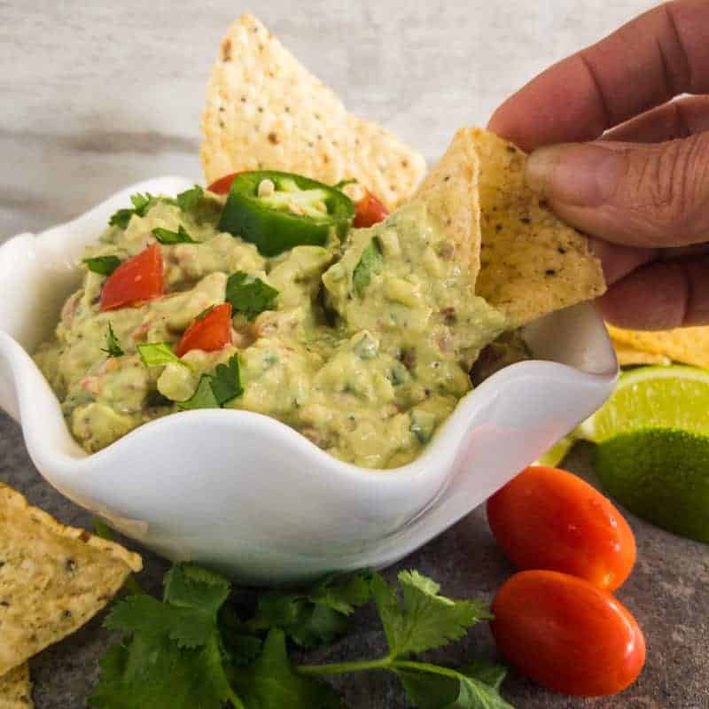 Easy Guacamole in a white bowl garnished with tomato, jalapeno, lime, cilantro; with a had dipping a corn chip.