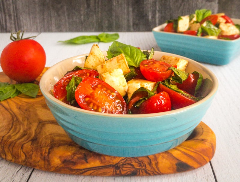 Caprese Salad in a blue bowl on top of a wood cutting board with a tomato and basil in the background.