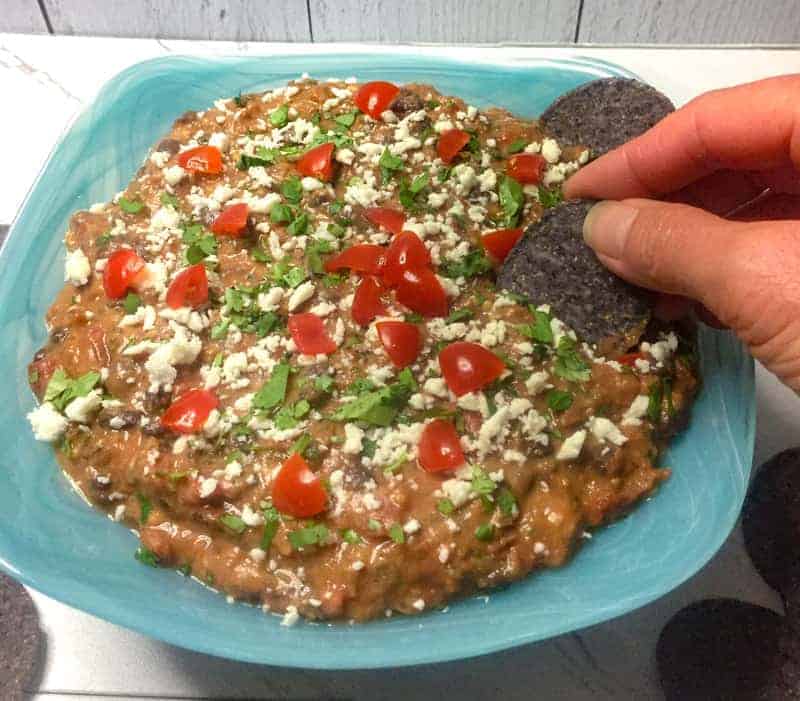 Fiesta Black Bean Dip in a blue bowl with fingers holding a chip.