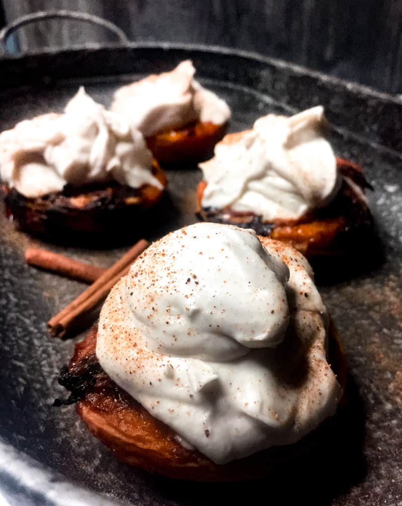 Grilled peaches, topped with coconute whipped cream on a glavinized tray with cinnamon sticks for garnish.