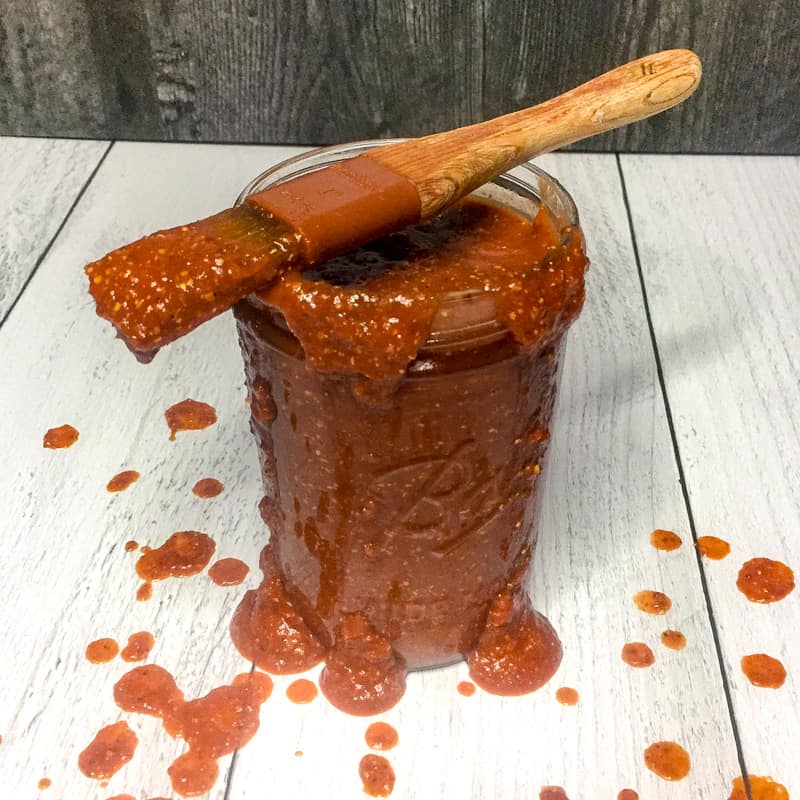 Jar of Homemade Barbecue Sauce on light background with a basting brush on top, surrounded by drips of sauce