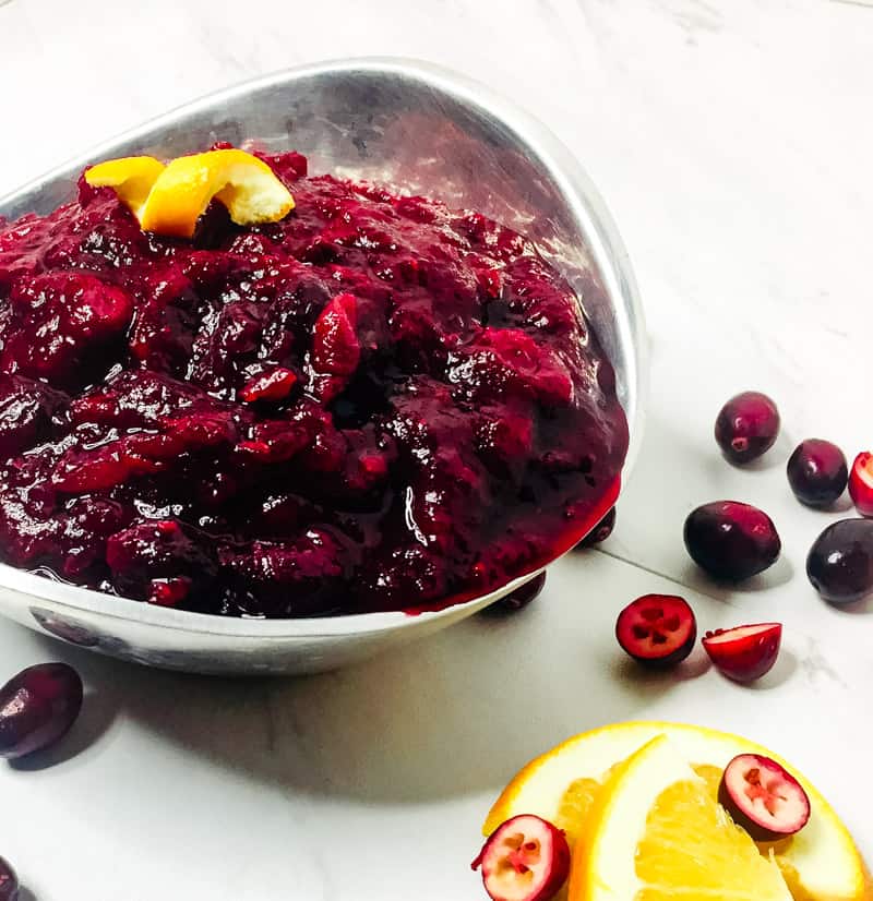 Three quarter side shot of Homemade Spiced Cranberry Sauce in a silver dish garnished with an orange peel with cranberries and orange slices surrounding the bowl.