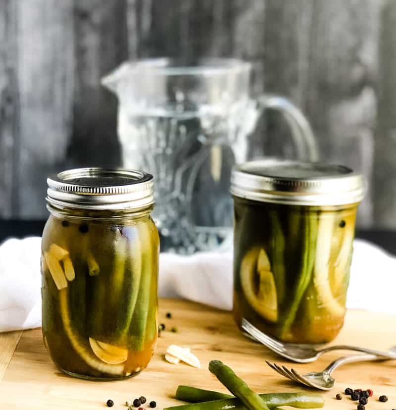 Two jars of Quick Pickled Green Beans on a wood cutting board with beans, peppercorns, and sliced garlic scattered.