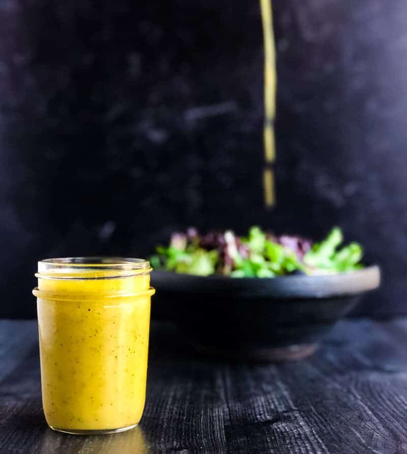 Jar of Creamy Lemon Honey Dijon Vinaigrette in foreground with mixed green salad in the background while a drizzle of dressing rains on the salad.