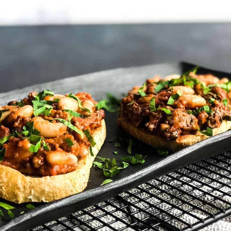 Beans and Sausage on crusty sourdough bread on black platter garnished with parsley.