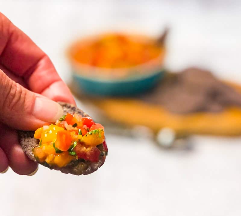 Close up of hand holding a blue corn chip with mango salsa; blurred salsa bowl in background.