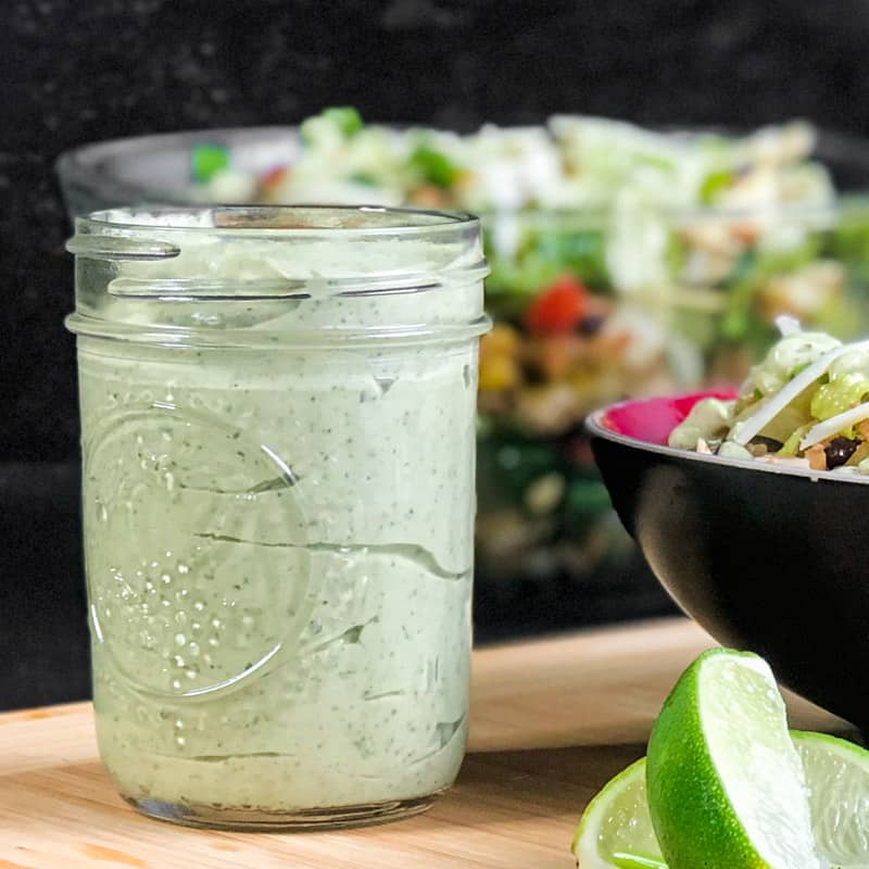 Close up of Avocado Crema in a mason jar with taco salad blurred in the background and sliced limes in the foreground.
