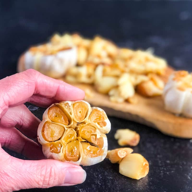 Close up of a hand holding a head of roasted garlic over a cutting board.