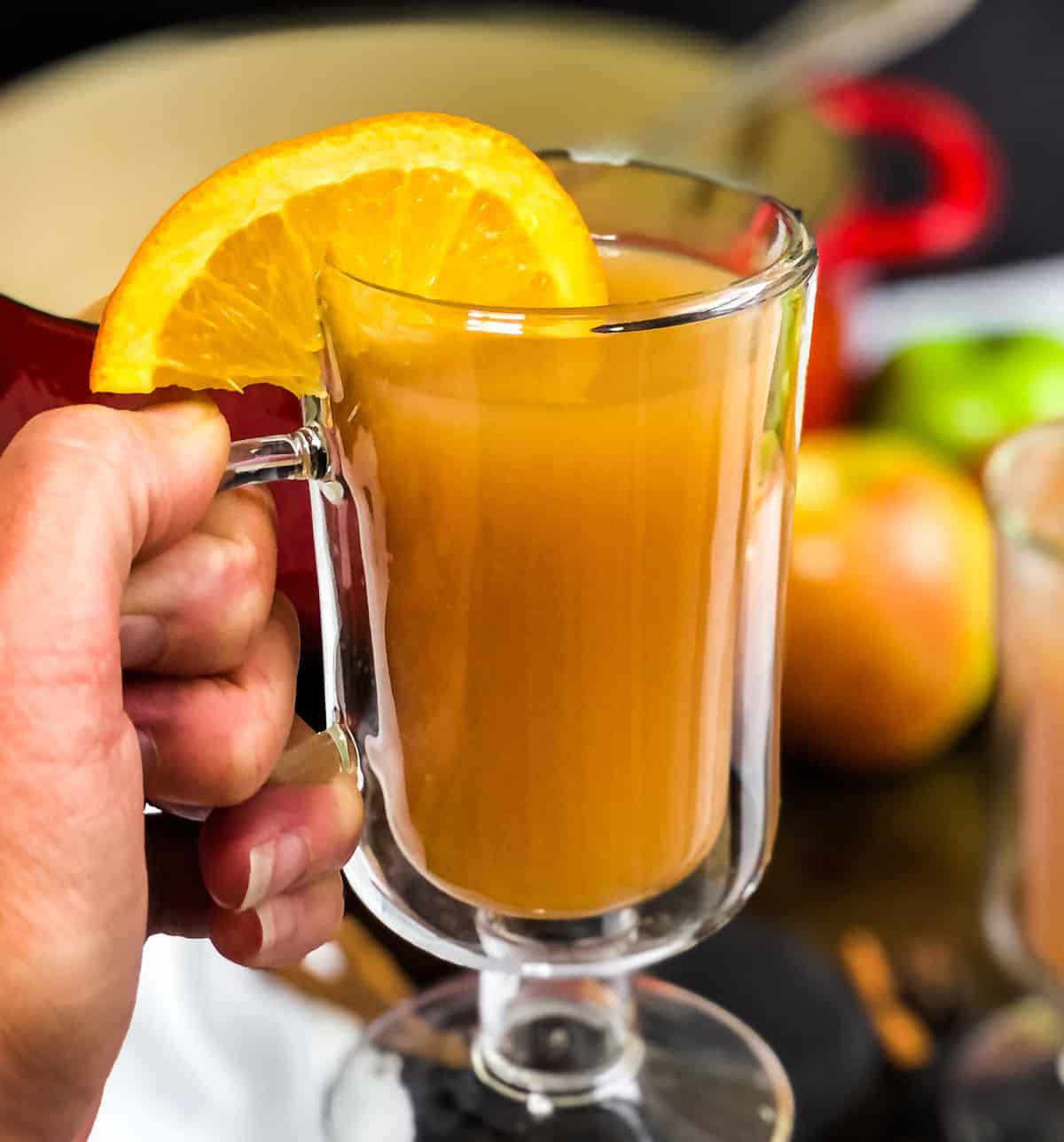 Hand holding a glass mug of spiced apple cider garnished with an orange slice.