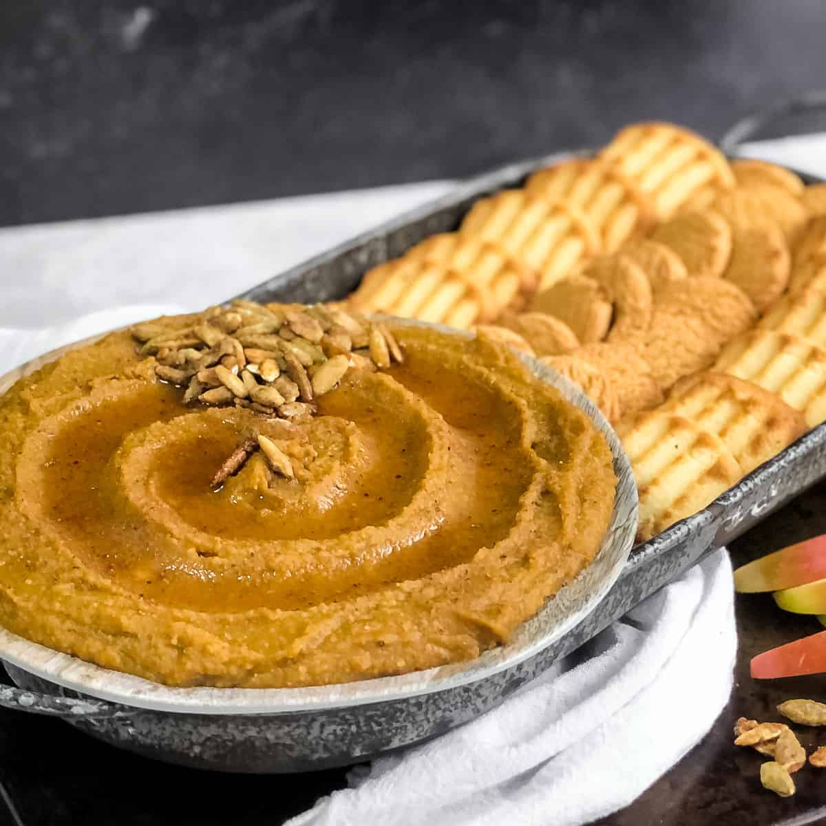 Pumpkin Pie Hummus swirled on a plate drizzled with maple syrup and garnished on the side with cookies and apples for dipping.