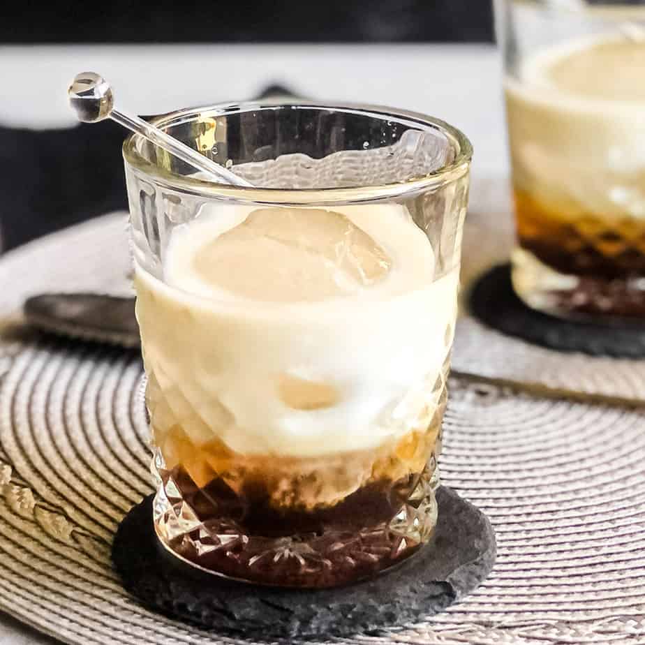 Peanut Butter White Russian in a rocks glass on a black coaster with another cocktail blurred. 