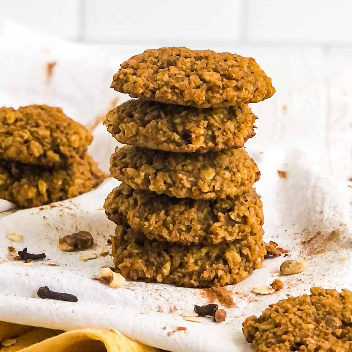 Stack of five Pumpkin Oatmeal Cookies on a white tea towel garnished with walnut pieces, whole cloves, and sprinkles of cinnamon.